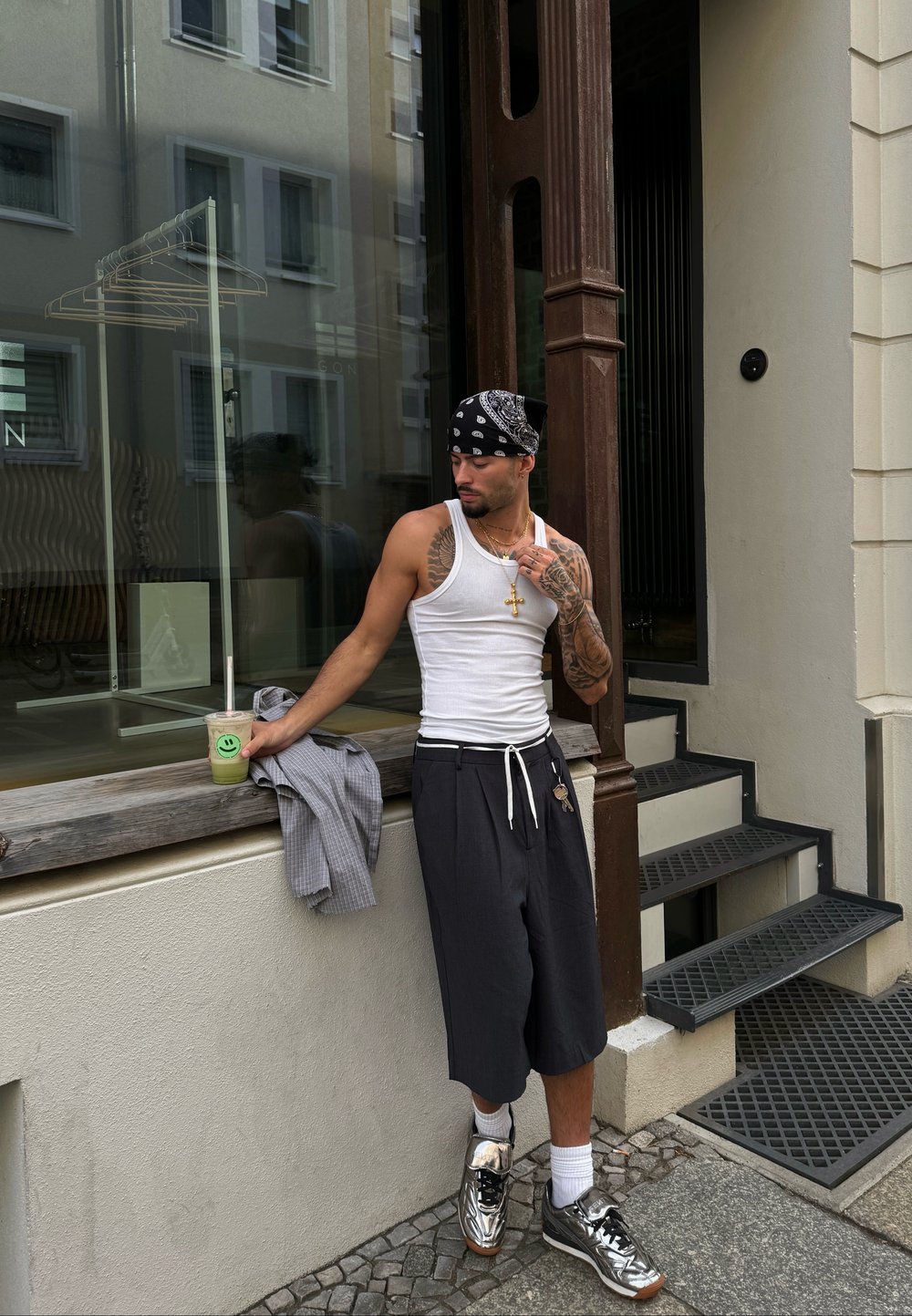 Débardeur blanc, short gris, baskets argentées. Tiens une boisson avec un visage souriant. Tatouages visibles sur le bras. Debout à côté d'une fenêtre.