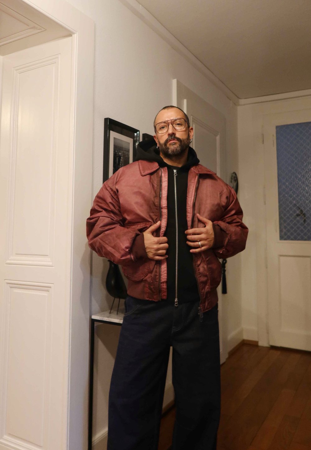 Bordo bomber jaka ar spīdīgu virsmu, priekšpusē ar rāvējslēdzēju un oversize piegriezumu, kombinēta ar tumšiem džinsa biksēm un melnu kapuci.