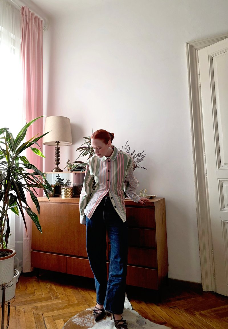 Striped button-up shirt with green, pink, and white hues; dark jeans; standing on a rug, surrounded by plants and wooden furniture.