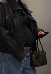 Veste en cuir noire avec fermetures éclair et boutons-pression, haut noir, jean taille haute bleu, tenant des lunettes de soleil foncées et un petit sac à main marron.