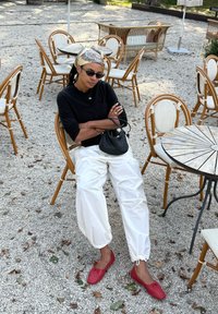 Γυναίκα που φοράει μαύρο τοπ, λευκό παντελόνι, κόκκινες μπαλαρίνες και γυαλιά ηλίου κάθεται με σταυρωμένα τα χέρια σε υπαίθριο καφέ με ψάθινες καρέκλες και χαλίκι κάτω.