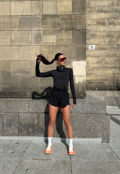 Camisa negra de manga larga y pantalones cortos, combinados con zapatillas deportivas blancas y naranjas. Unas gafas de sol de gran tamaño y una bufanda negra con estampado completan el conjunto.