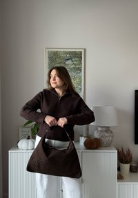 Brązowa pikowana torebka o miękkiej fakturze i pojedynczym uchwycie, trzymana na tle neutralnej dekoracji i roślin domowych.