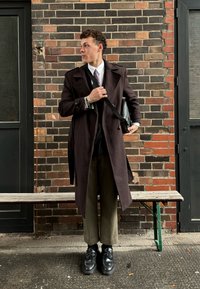 Μακρύ καφέ παλτό πάνω από ένα μαύρο κοστούμι, γκρι γραβάτα, ελιές πράσινα παντελόνια και μαύρα παπούτσια. Κρατά μια μαύρη τσάντα, στέκεται δίπλα σε έναν τοίχο από τούβλα.