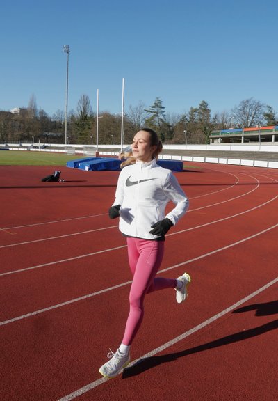 Corredora con cortavientos blanco con acentos negros, mallas rosas y zapatillas blancas. Superficie de pista roja y cielo azul claro al fondo.