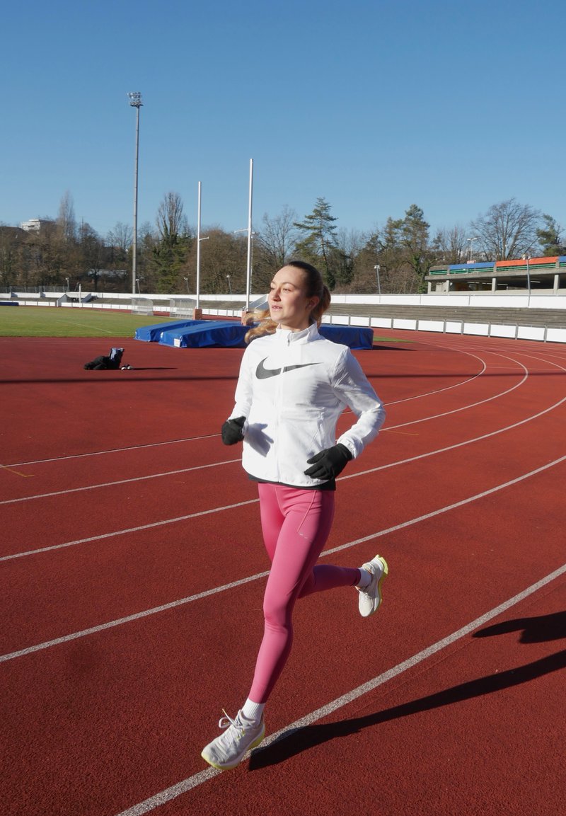 Coureuse en veste coupe-vent blanche avec des accents noirs, leggings roses et baskets blanches. Surface de piste rouge et ciel bleu clair en arrière-plan.