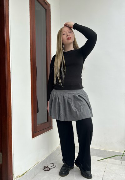 Top negro de manga larga acanalado combinado con una falda gris con pliegues sobre pantalones anchos negros y zapatos negros. Entorno interior con una ventana.