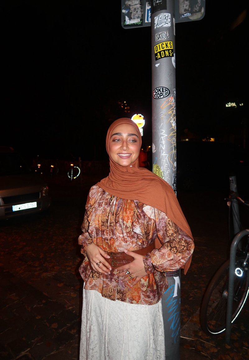 Eine Person trägt einen braunen Hijab und eine mehrfarbige, gemusterte Bluse mit Rüschenärmeln, die mit einem braunen Gürtel in der Taille zusammengezogen ist, und steht neben einem Pfosten.