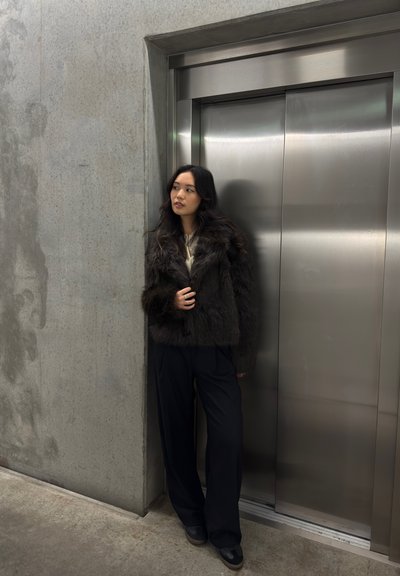 Mujer con cabello largo y oscuro, vestida con una chaqueta de piel y pantalones negros, apoyada contra la pared junto a las puertas del ascensor de acero inoxidable cerradas.