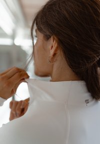 Hvit treningsskjorte med høy krage, glatt tekstur og synlige sømdetaljer. Brukeren justerer kragen, noe som avdekker en minimalistisk logo på baksiden.
