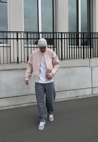 Hombre con chaqueta rosa, pantalones grises, zapatillas blancas y gorro gris caminando por la acera cerca de un edificio con grandes ventanas y barandilla negra.