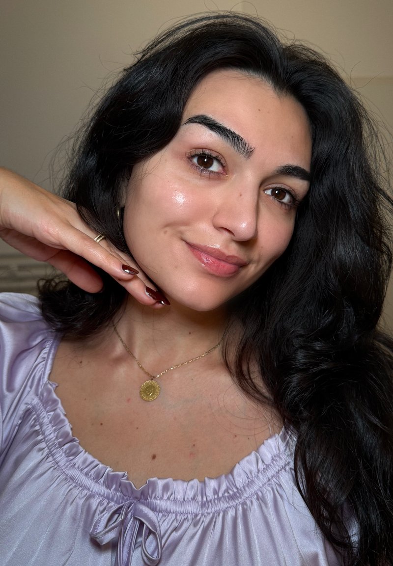 Portrait of a person with long, dark hair, wearing a lavender blouse with gathered neckline. A gold pendant necklace and polished nails are visible.