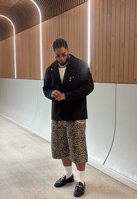 Černý blazer přes bílé triko, doplněný leopardími kraťasy. Černé loafers a bílé ponožky dotvářejí outfit, který zahrnuje vrstvené šperky.