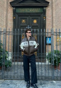 Hnedý a krémový kožený bomber jaket s textom "MOTOR SPORT", čierne nohavice a slnečné okuliare, stojaci pred bránou kostola.