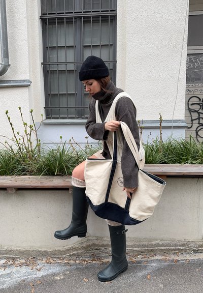 Bolsa de lona beige con detalles en azul marino, diseño grande, asas resistentes y un logo. El modelo lleva un gorro de lana negro, un suéter gris y botas de goma verdes.