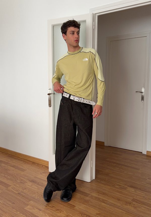 Jeune homme appuyé sur un cadre de porte blanc, portant une chemise jaune à manches longues, un pantalon large noir, une ceinture blanche et des chaussures noires dans une pièce avec un sol en bois.