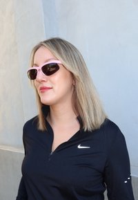 Mulher com cabelo loiro, usando óculos de sol desportivos cor-de-rosa e uma camisola preta com fecho da Nike, de pé contra uma parede de cor clara.
