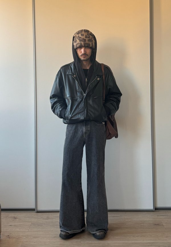 Homme portant un chapeau en fausse fourrure à imprimé léopard, une veste en cuir noire sur un sweat à capuche noir, un jean gris à jambes larges, des chaussures noires, et portant un sac à bandoulière marron.