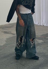 Baggy blue denim jeans featuring large ripped sections and frayed edges. They are paired with a black oversized sweatshirt and black shoes.