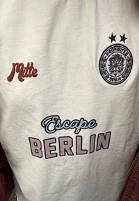 Bijela tkanina s ispisanim tekstom "Escape BERLIN," smeđi tekst "Mitte" s lijeve strane, i kružni crni emblem s dvije zvijezde i tekstom "Prohibited Berlin" s desne strane.