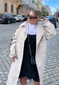 Trench beige claro con detalles negros, superpuesto sobre una blusa blanca bordada y un vestido negro. Accesorio con un pequeño bolso negro.