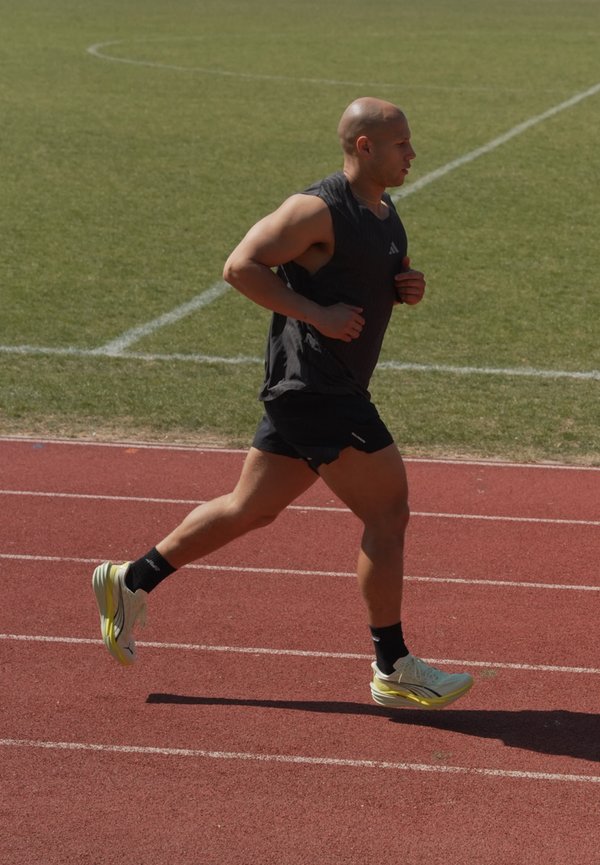 Uomo con abbigliamento sportivo nero che corre su una pista esterna rossa con un campo di erba verde sullo sfondo.
