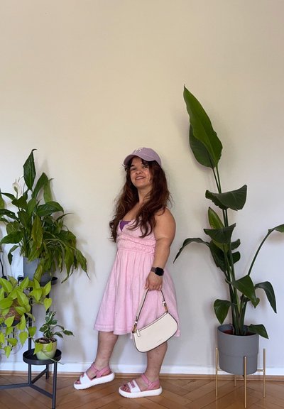 Vestido rosa claro, gorra púrpura, bolso blanco y sandalias rosas con suelas blancas. El fondo presenta plantas verdes y paredes de color claro.