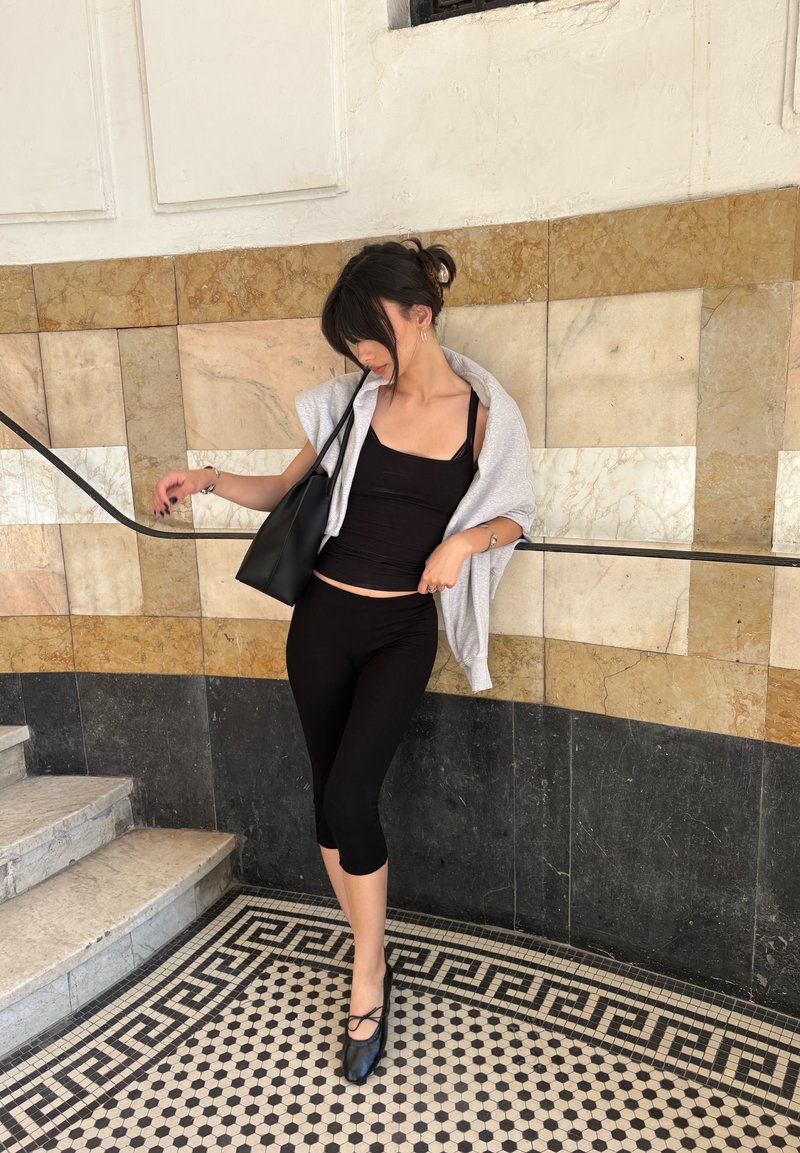 Jeune femme en tenue noire avec un pull gris drapé sur les épaules, appuyée sur la rampe près d'un mur carrelé et d'un sol en mosaïque près des escaliers.