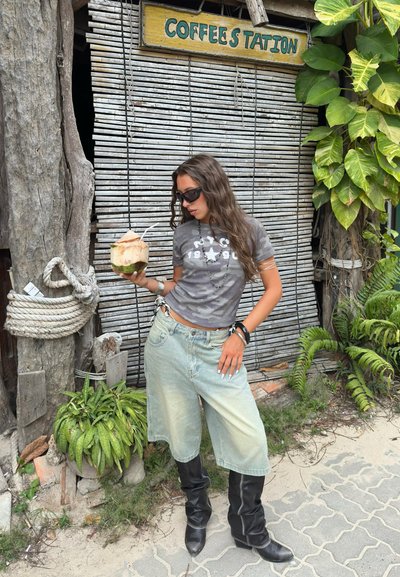 Joven mujer con gafas de sol, camiseta gris, shorts de mezclilla y botas negras sosteniendo un coco con pajilla cerca de un cartel de estación de café.