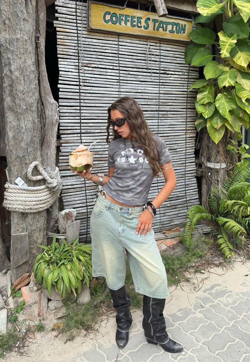 Νεαρή γυναίκα με γυαλιά ηλίου, γκρι μπλουζάκι, τζιν σορτς και μαύρες μπότες, κρατώντας ένα καρύδα με καλαμάκι κοντά σε πινακίδα καφετέριας.