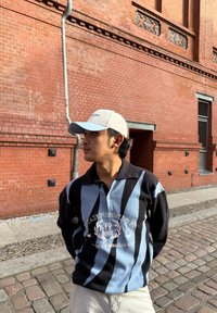 Musta- ja siniraidallinen poolopaita, jossa on kirjailtu logo, yhdistetty vaaleansinisellä lippalakilla. Taustalla on punatiiliseinä.