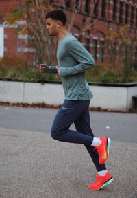 Homme athlétique courant, portant un t-shirt à manches longues turquoise et un pantalon bleu marine. Baskets orange vif et vertes avec des chaussettes blanches. Arrière-plan urbain.
