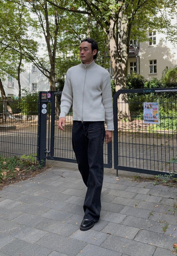 Hombre caminando por un camino pavimentado cerca de una puerta metálica en una zona sombreada del parque, con un suéter blanco con cremallera, pantalones negros y zapatos negros.