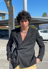 Blazer preto de duas fileiras com um ajuste sob medida, usado sobre uma camisa clara. Apresenta um colar de corrente prateada. O fundo inclui um estacionamento e árvores.