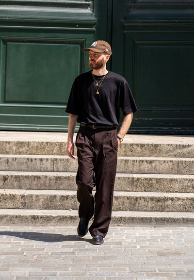 Camiseta negra de gran tamaño, pantalones de vestir marrones con pliegues, zapatos negros y una gorra marrón. Los accesorios incluyen cadenas de oro en capas. Fondo verde.