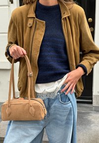 Sac à main en daim beige avec poignées courtes, forme rectangulaire, fermeture éclair, associé à une veste en daim marron et un pull bleu superposé.