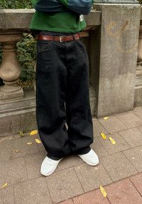 Svart baggy bukser laget av denim, sammen med et brunt skinnbelte. Hvite joggesko synlig, stående på et pavebelagt underlag med falt blader.