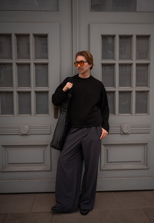 Joven con pantalones oscuros y holgados, camisa negra de manga larga y gafas de sol naranjas, está apoyado en una puerta gris sosteniendo un bolso de hombro negro.