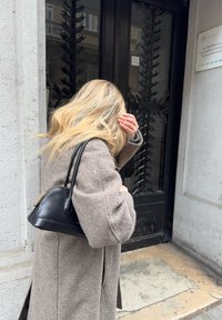 Cappotto di lana marrone con maniche ampie, che porta una piccola borsa strutturata nera, di fronte a una porta in metallo scuro con elementi decorativi.