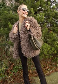 Manteau marron à poil long en fausse fourrure, pantalon noir ajusté, accompagné d'un sac seau vert foncé et de lunettes de soleil, entouré de verdure.