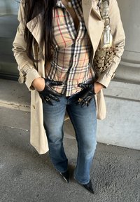 Trench beige, camicia a quadri color sabbia e rosso, guanti di pelle neri, jeans blu a zampa, e décolleté nere con punta; borsa marrone con motivo.