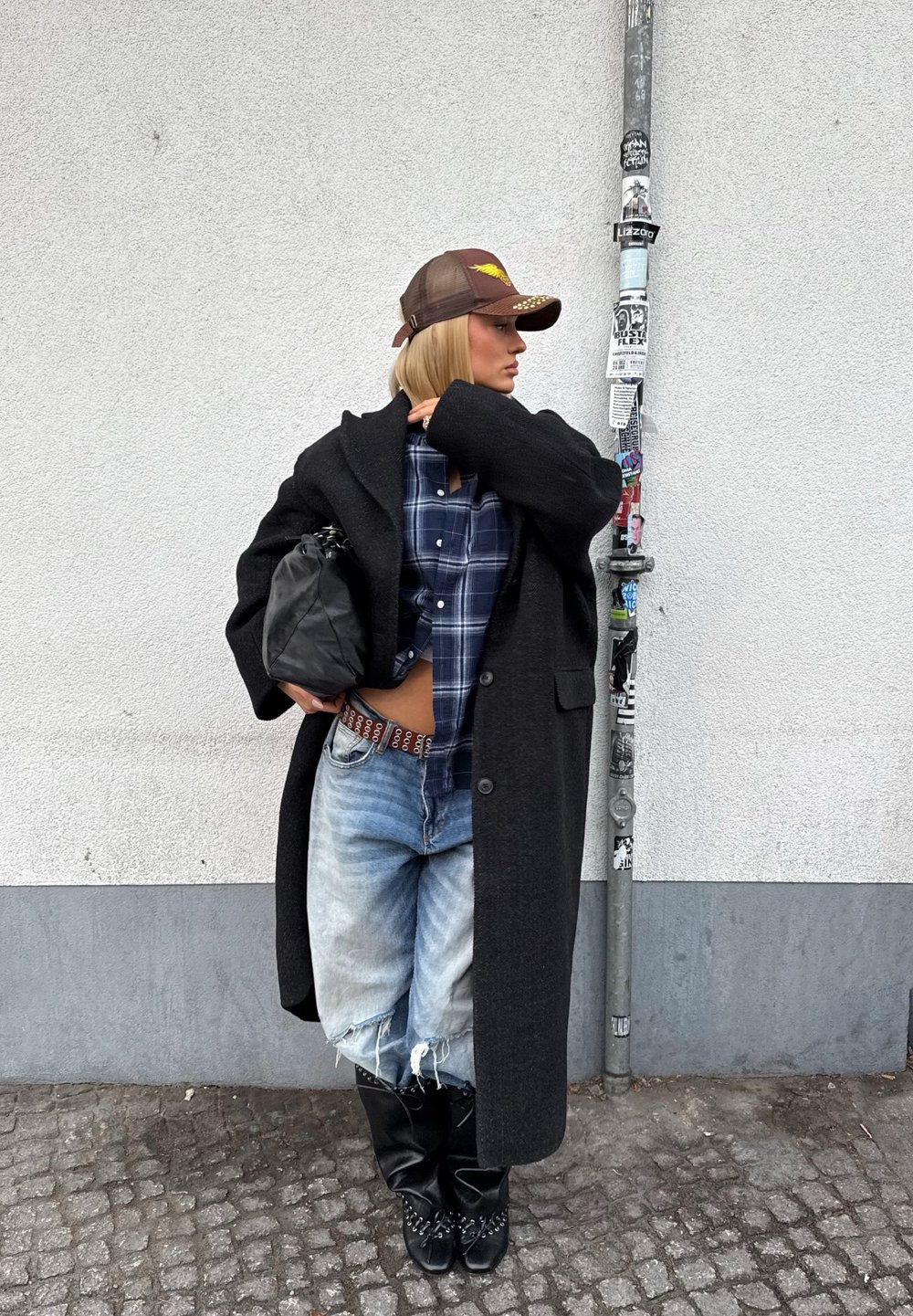Femme portant un manteau noir surdimensionné, une chemise à carreaux, un jean déchiré, des bottes noires et une casquette marron, debout contre un mur blanc avec un poteau couvert d'autocollants.