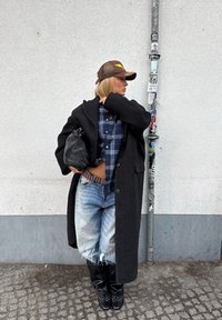 Femme portant un manteau noir surdimensionné, une chemise à carreaux, un jean déchiré, des bottes noires et une casquette marron, debout contre un mur blanc avec un poteau couvert d'autocollants.