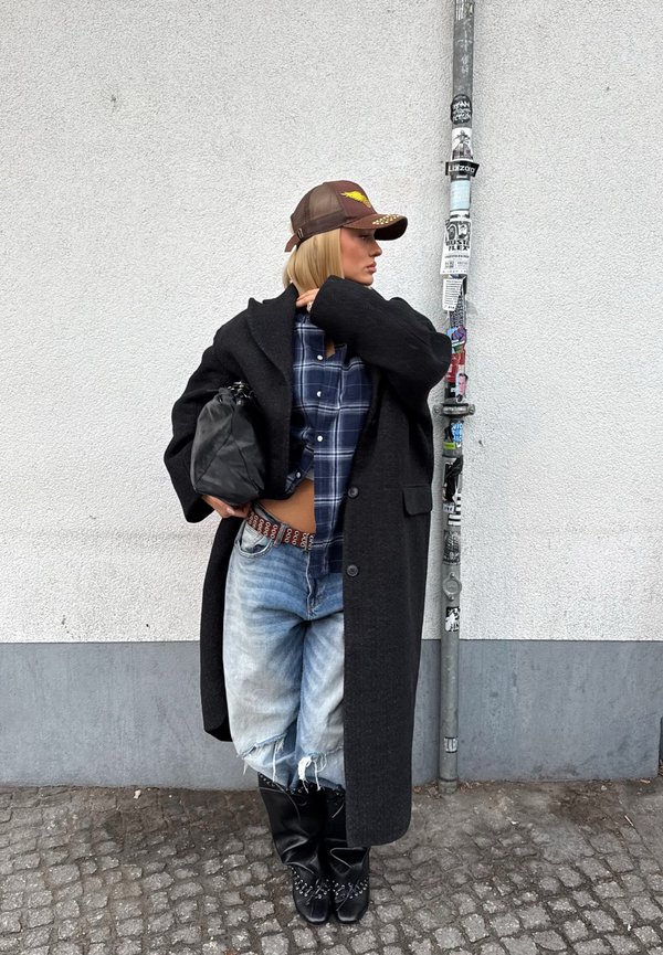 Vrouw in een oversized zwarte jas, geruit overhemd, gescheurde spijkerbroek, zwarte laarzen en een bruine pet, staand tegen een witte muur met een paal bedekt met stickers.