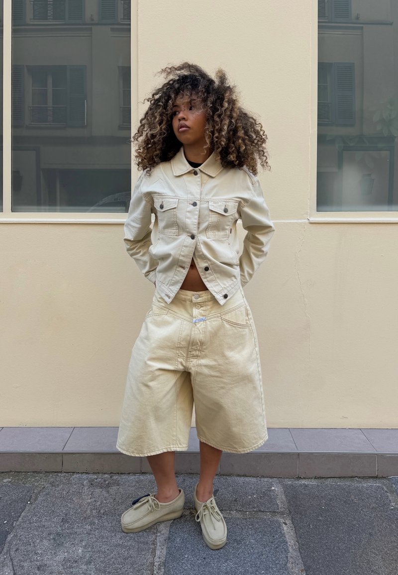 Femme aux cheveux bouclés debout, les mains derrière le dos, portant une veste crème claire, un short large assorti et des chaussures beiges devant un mur pâle.