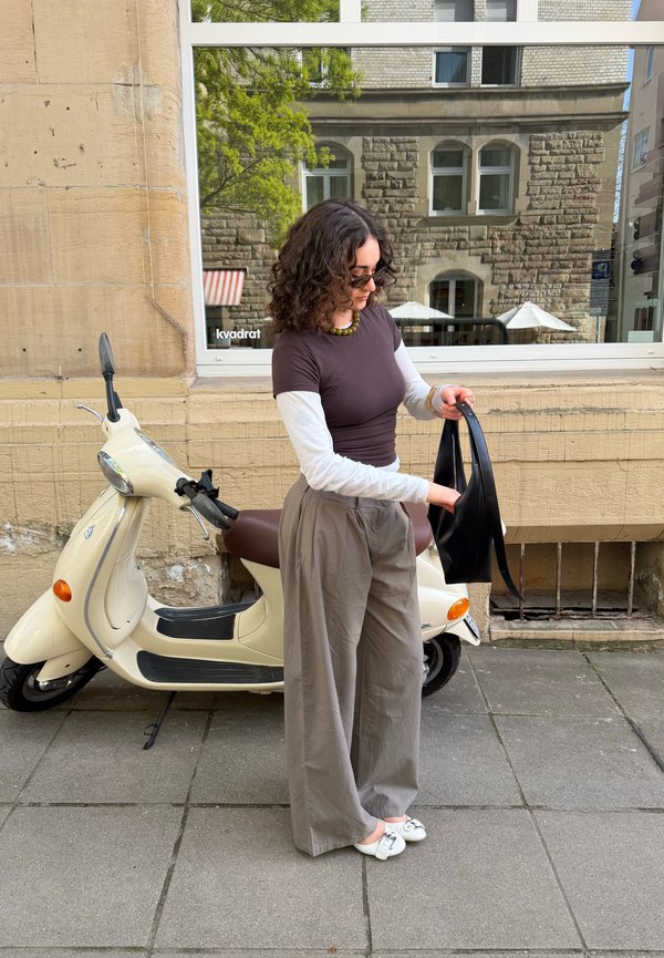 Vrouw met krullend haar die een zonnebril en gelaagde tops draagt, staat naast een crèmekleurige scooter op een stadsstoep en houdt een zwarte tas vast.
