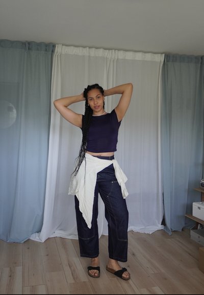 Top corto de color púrpura oscuro combinado con pantalones de mezclilla anchos en un tono oscuro, un cinturón blanco alrededor de la cintura y sandalias negras tipo deslizante. Fondo de suelo de madera.