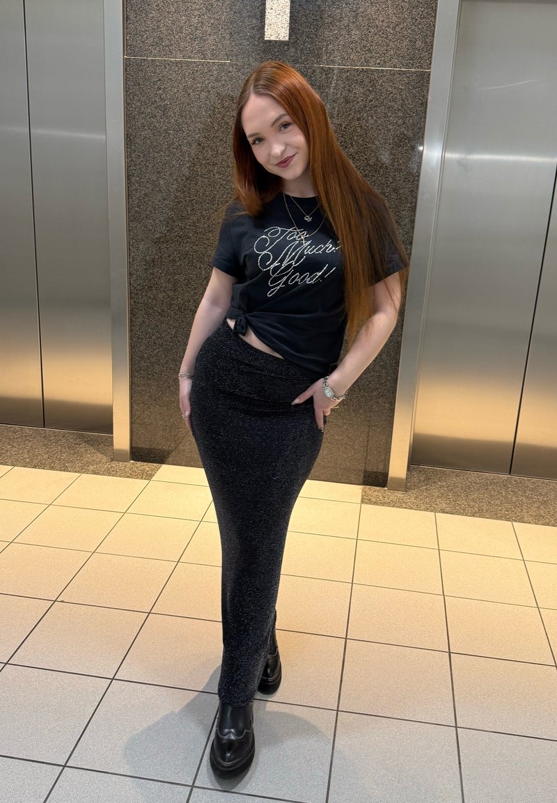 Jeune femme aux longs cheveux roux portant un t-shirt noir graphique et une jupe ajustée scintillante, debout entre deux portes d'ascenseur dans un couloir carrelé.