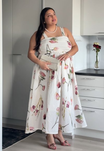 Mujer con un vestido blanco con estampado floral, collar y pulsera de perlas, sostiene un pequeño bolso beige, de pie en una cocina moderna.