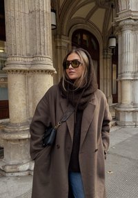 Cappotto in lana marrone con vestibilità oversize, indossato sopra un maglione scuro, abbinato a occhiali da sole e una piccola borsa nera; colonne di pietra testurizzate sullo sfondo.